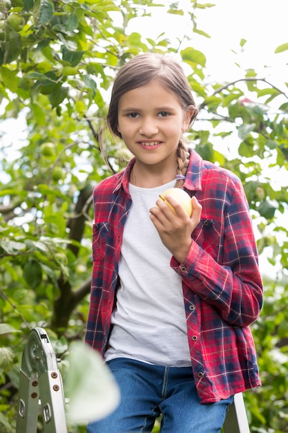 Zbliżenie portret pięknej uśmiechniętej dziewczyny w kraciastej koszuli pozowanie w ogrodzie i gryzienie jabłka