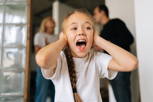 Zbliżenie portret podekscytowanej zdenerwowanej córeczki krzyczącej, odwracającej wzrok, zakrywającej uszy, podczas gdy wściekli rodzice kłócą się i walczą na tle w domu