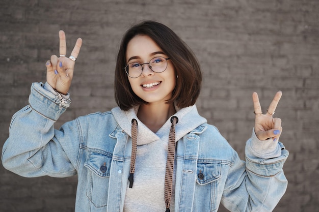 Zbliżenie Portret Queer Słodkie Dziewczyny W Okularach Studentka Pokazując Kawaii Gest Pokoju I Uśmiechnięta Kamera Ciesząc Się Beztroskim życiem Uniwersytetu Młodzieżowego Stojącego W Pobliżu Ceglanego Muru Na Ulicy