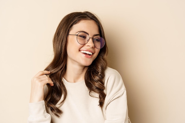 Zbliżenie portret stylowej brunetki w okularach, śmiejącej się i uśmiechniętej, pozującej w okularach na beżowym tle