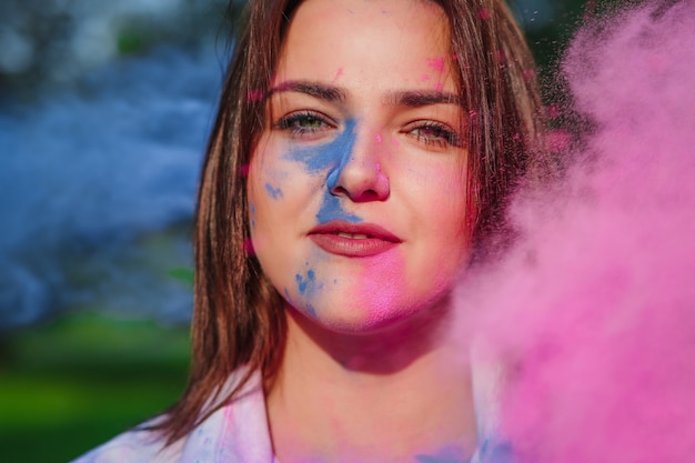 Zbliżenie portret uroczej brunetki pozowanie w chmurze różowej i niebieskiej suchej farby Holi