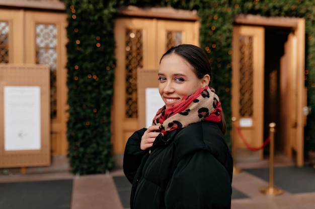 Zbliżenie portret wesoły kaukaski kobieta z jasnym makijażem dotykając jej szalik na rozmycie tła Zdjęcie modnej dziewczyny z pięknymi brązowymi włosami uśmiecha się do kamery