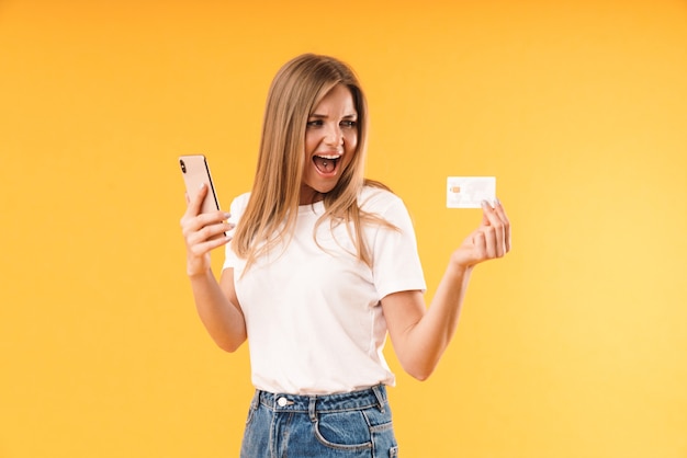 Zbliżenie portreta wesołej blond kobiety noszącej zwykłą koszulkę krzyczącą, trzymając smartfon i kartę kredytową na białym tle nad żółtą ścianą