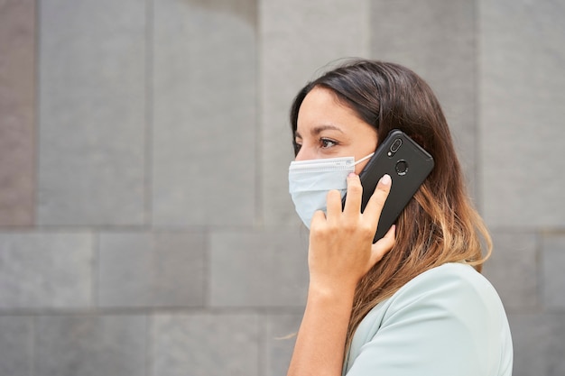 Zbliżenie Pracującej Kobiety Z Maską Rozmawia Przez Telefon. Po Lewej Stronie Znajduje Się Puste Miejsce Na Integrację Tekstu. Tło To Szara ściana.