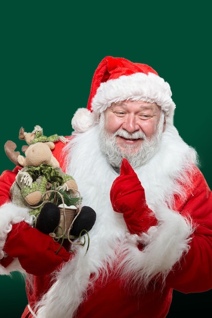 Zbliżenie przód portret szczęśliwy Santa Claus ubrany w czerwony płaszcz, rękawiczki i kapelusz, trzymając w ręce zabawkę, wskazując palcem wskazującym, na białym tle zielony.