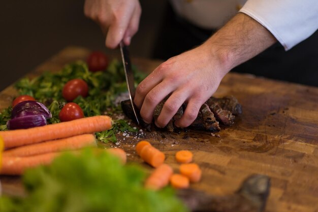 zbliżenie rąk szefa kuchni w kuchni hotelowej lub restauracyjnej przygotowującej stek wołowy z dekoracją warzywną