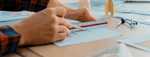 Zbliżenie ręki inżyniera architekta używającego linijki do dojrzewania i narysowania planu na stole konferencyjnym drewnianym ołówkiem i rozproszonym planem w nowoczesnym biurze architektonicznym