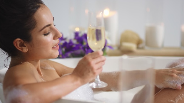 Zbliżenie seksowna kobieta myje nogi pianką w domowej łazience Zmysłowa dziewczyna relaksuje przy lampce wina w wannie Brunetka pije szampana w luksusowej kąpieli