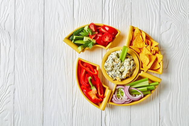 Zdjęcie zbliżenie: ser szpinakowy karczoch liściasty dip z chipsami tortilla, ogórkiem, selera naciowego, plastrami pomidora i czerwoną papryką, widok poziomy z góry, układ płaski, wolna przestrzeń