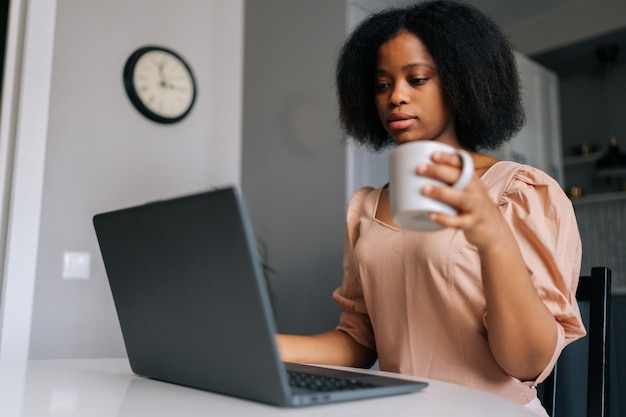 Zbliżenie skoncentrowanej Afroamerykanki, freelancerki, piszącej na laptopie, patrzącej na ekran urządzenia i pijącej kawę lub herbatę rano, siedzącej przy stole