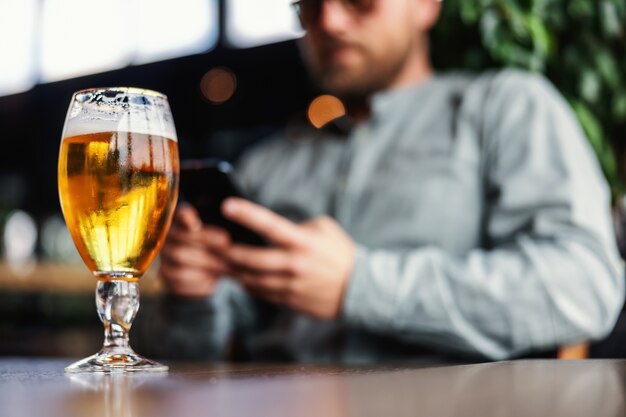 Zbliżenie świeżego zimnego piwa w szkle. W tle mężczyzna pisze wiadomość przez telefon.