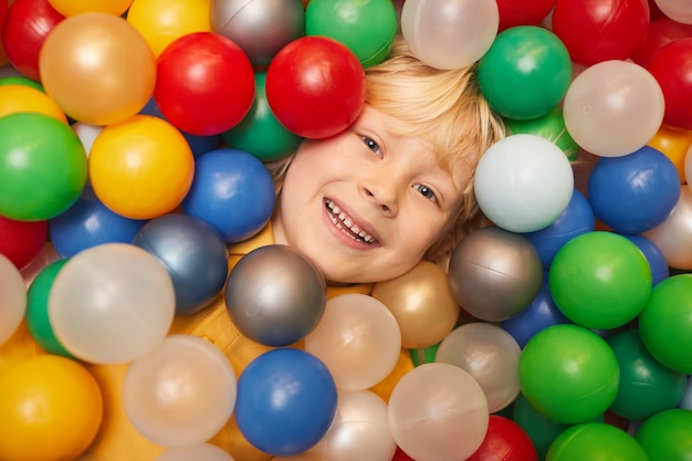 Zdjęcie zbliżenie: szczęśliwy chłopiec o blond włosach, uśmiechając się z przodu, leżąc wśród kolorowych kulek i grając