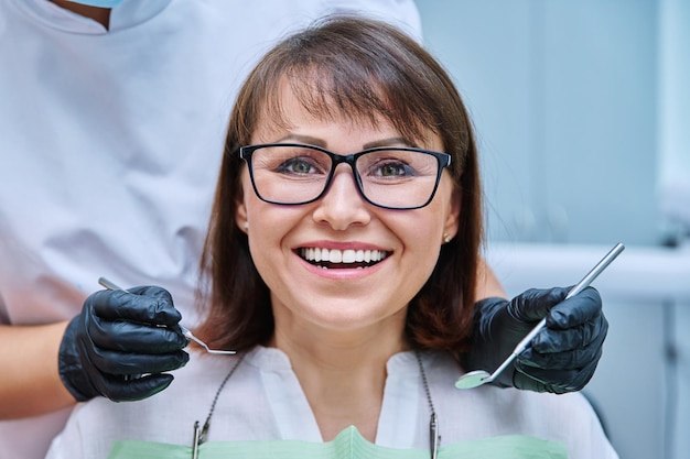 Zbliżenie twarzy uśmiechniętej dojrzałej kobiety z zębami w stomatologii z rękami z narzędziami do badania dentysty Leczenie protetyka implantacja wybielanie higiena koncepcja opieki zdrowotnej stomatologia