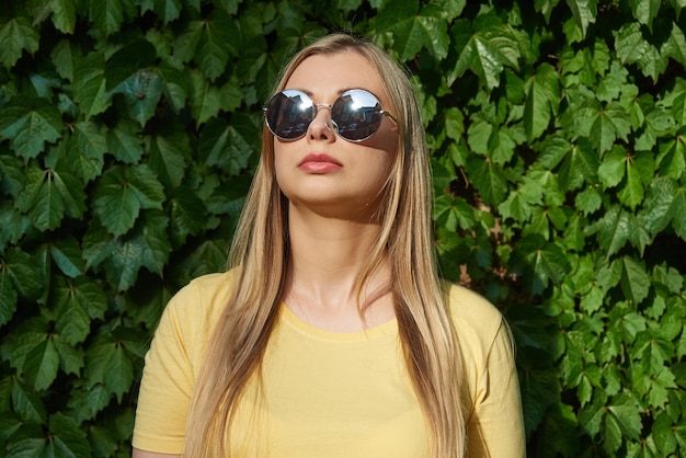 Zbliżenie uroczej kobiety w okularach przeciwsłonecznych stoi na tle zielonego, naturalnego ulistnienia b