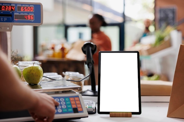 Zbliżenie urządzenia cyfrowego wyświetlającego izolowany szablon makiety w sklepie spożywczym przyjaznym dla środowiska Telefon tablet z pustym wyświetlatłem makiety umieszczonym w pobliżu elektronicznej skali pomiarowej używanej przez sprzedawcę