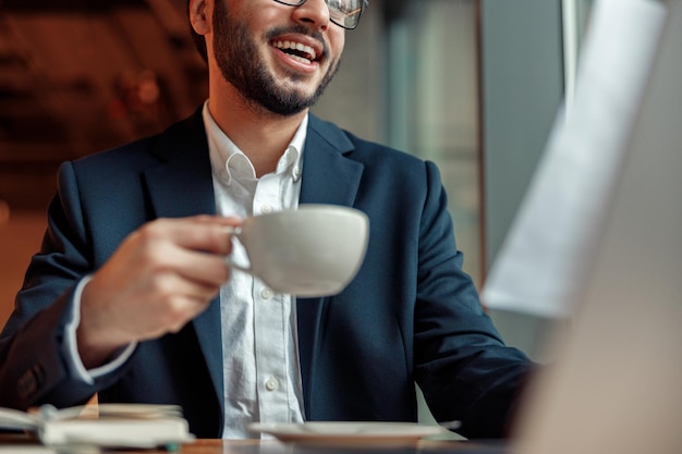 Zbliżenie uśmiechniętego nierozpoznawalnego biznesmena pijącego kawę i pracującego laptopa w kawiarni