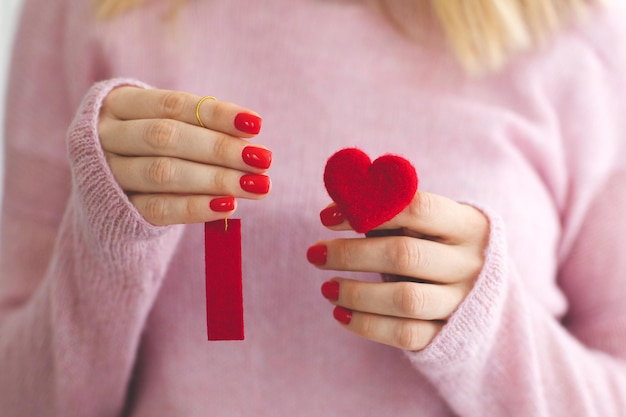 Zbliżenie womans ręce czerwony manicure trzyma serce pod wełną pastelowy różowy sweter z dzianiny tle