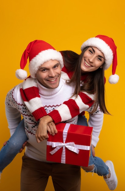 Zbliżenie zdjęcie pięknej podekscytowanej młodej pary w świątecznych czapkach Mikołaja, podczas gdy facet trzyma swoją dziewczynę z pudełkiem na plecach
