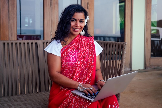Zdalna koncepcja pracy wymarzona praca indyjska kobieta biznesu w czerwieni stylowe sari kręcone fryzurytoothy white uśmiech i kwiat we włosach pracując z laptopem siedząc w letniej kawiarni nad morzem