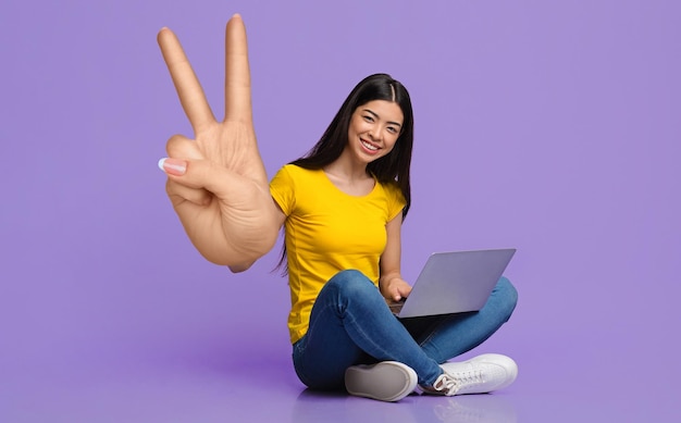 Zdalna praca lub edukacja Uśmiechnięta młoda Azjatka siedząca z laptopem pokazująca duży gest pokoju lub zwycięstwa