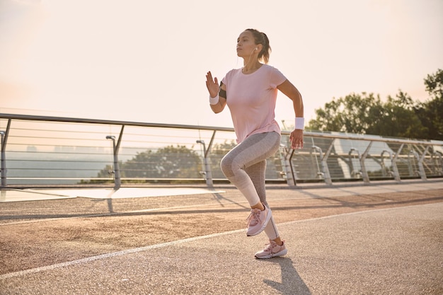 Zdecydowanie silna atletyczna latynoska sportsmenka w średnim wieku w sportowej stroju podczas porannego biegu na bieżni wzdłuż miejskiego mostu z piękną przyrodą w tle