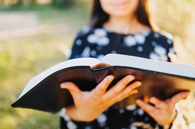 Zdekoncentrowana młoda kobieta trzyma i czyta swoją Biblię, na zewnątrz z podświetleniem słońca. Duchowe odrodzenie