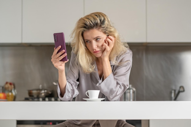 Zdenerwowana dziewczyna z telefonem pijąca wino w kuchennym poranku ładnej kobiety smutnej damy używającej smartfona soc
