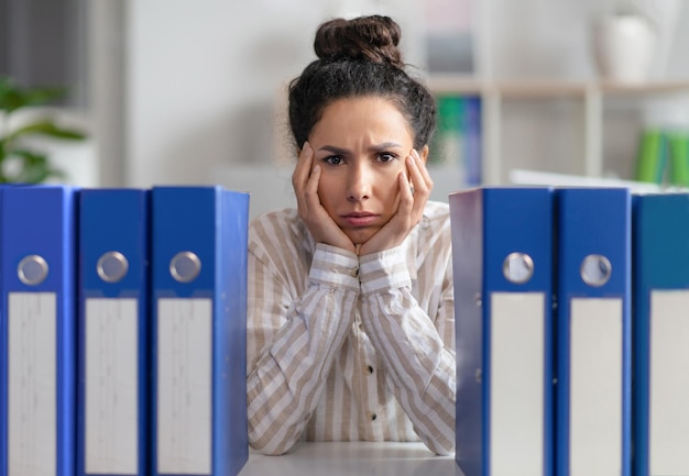 Zdenerwowana kobieta bizneswoman siedząca w miejscu pracy ze stosem folderów i patrząca na aparat fotograficzny