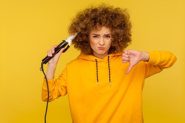 Zdenerwowana niezadowolona kobieta używa lokówki do robienia fryzury afro i pokazuje kciuk w dół niechęć do prasowania loków wyrażając niezadowolenie z narzędzia zła fryzura w studio strzał na białym tle