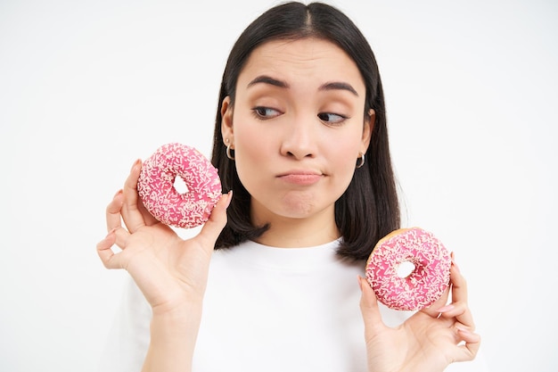 Zdenerwowana szlochająca dziewczyna nie może jeść fast foodów pokazuje pyszne przeszklone pączki chce ugryźć białe tło