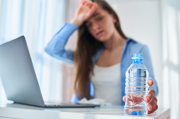 Zdenerwowana zmęczona pracująca kobieta cierpiąca na upał, pragnienie i upały ochładza się butelką z zimną wodą podczas pracy online przy komputerze w ciepły letni dzień