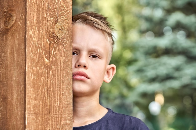 Zdenerwowany blond chłopiec w ciemnej koszulce chowa się za drewnianym płotem w zielonym słonecznym letnim parku miejskim