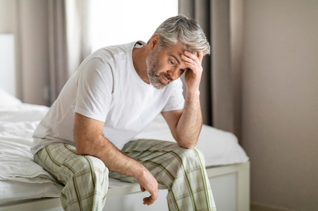Zdenerwowany mężczyzna w średnim wieku siedzi sam na łóżku w domu