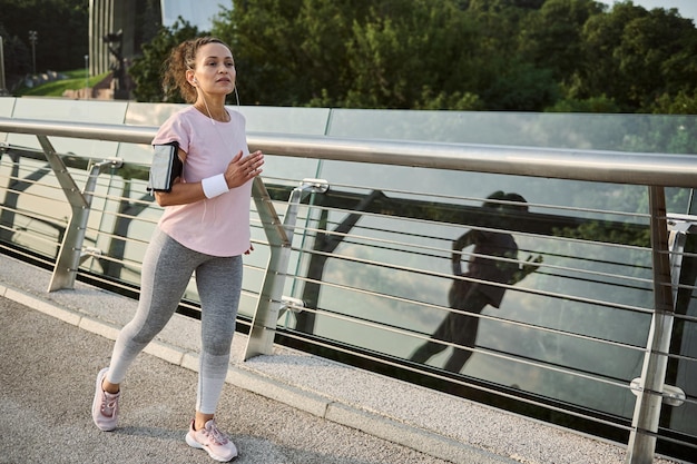 Zdeterminowana, Silna, Wysportowana Kobieta Uprawiająca Jogging Afroamerykanka W średnim Wieku W Stroju Sportowym Podczas Porannego Biegu, Biegająca Na Bieżni Wzdłuż Miejskiego Mostu Z Piękną Przyrodą W Tle