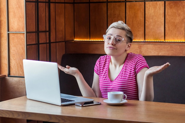 Zdezorientowana młoda dziewczyna freelancer z blond krótkimi włosami w różowej koszulce i okularach siedzi w kawiarni i pracuje na laptopie z uniesionymi rękami i zdziwioną twarzą, patrząc na kamerę w pomieszczeniu