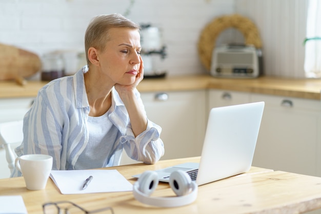 Zdezorientowana starsza kobieta trzymająca głowę w dłoniach i patrząca na laptopa podczas pracy online w domu