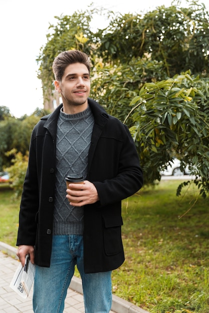 Zdjęcie atrakcyjnego mężczyzny w wieku 30 lat w kurtce trzymającego papierowy kubek kawy na wynos podczas spaceru po zielonym parku