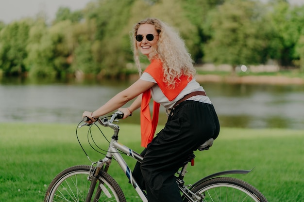 Zdjęcie atrakcyjnej młodej kobiety z kręconymi włosami jeździ na rowerze nosi okulary przeciwsłoneczne koszulka spodnie bluza na ramionach cieszy się letnim czasem świeżym powietrzem na świeżym powietrzu aktywnie spędza wolny czas z przyjaciółmi