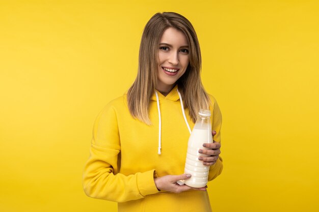 Zdjęcie atrakcyjnej pani trzymającej butelkę mleka, dobrze czuje się w produktach mlecznych. Nosi dorywczo żółtą bluzę z kapturem, izolowane żółte tło.