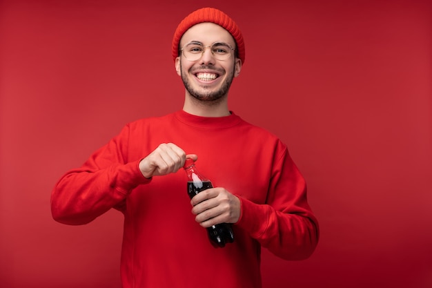 Zdjęcie atrakcyjny mężczyzna z brodą w okularach i czerwoną odzież. Szczęśliwy człowiek trzyma napój i otwiera bootle, na białym tle nad czerwonym tle.