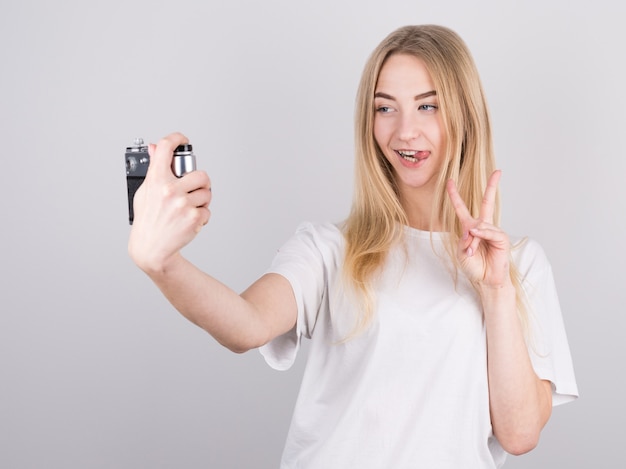 Zdjęcie blondynki w dorywczo t-shirt pokazujący znak pokoju i biorąc selfie ze starym aparatem