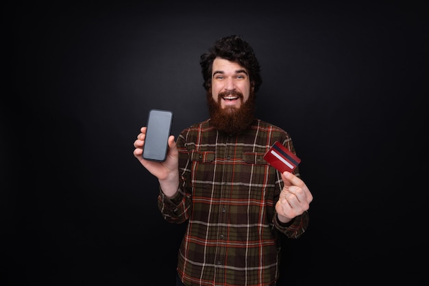 Zdjęcie brodatego faceta w koszuli, pokazującego smartfon i kartę kredytową, stojącego nad ciemną ścianą