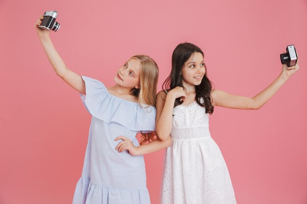 Zdjęcie Brunetki I Blondynki Urocze Dziewczyny Ubrane W Sukienki, Uśmiechając Się I Robiąc Zdjęcia Selfie Na Aparatach Retro, Na Białym Tle Na Różowym Tle