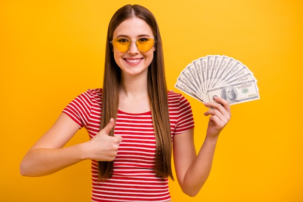 Zdjęcie całkiem ładna młoda dziewczyna trzymaj dolary gotówki wentylator zatwierdzający biorąc kredyt niezawodne wsparcie bankowe biznes nosić okulary przeciwsłoneczne w paski biała czerwona koszula żywy żółty kolor tła