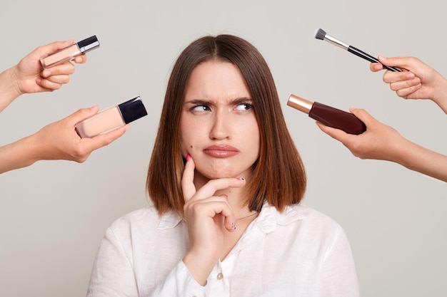 Zdjęcie dłoni kilku kosmetyczek trzymających swój sprzęt, które robią makijaż atrakcyjnej kobiecie stojącej z zamyśloną, zamyśloną miną
