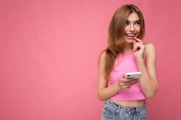 Zdjęcie dobrze wyglądającej młodej kobiety blondynka ubrana w różowy top pozowanie na białym tle na różowym tle z pustą przestrzenią trzymającą w ręku i używającą wiadomości sms z telefonu komórkowego, patrząc z boku
