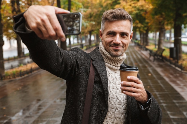 Zdjęcie Dojrzałego Mężczyzny Lat 30. W Ciepłych Ubraniach Spacerującego Po Jesiennym Parku Na świeżym Powietrzu I Robienia Selfie Na Telefonie Komórkowym