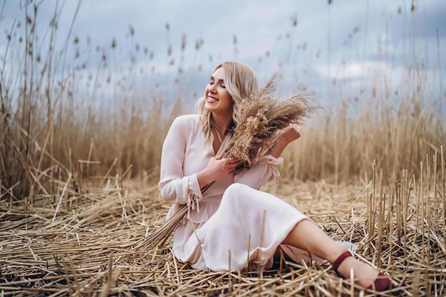 Zdjęcie dość uśmiechnięte dziewczyny z długimi blond kręconymi włosami w świetle długie drees siedzi w polu trzciny