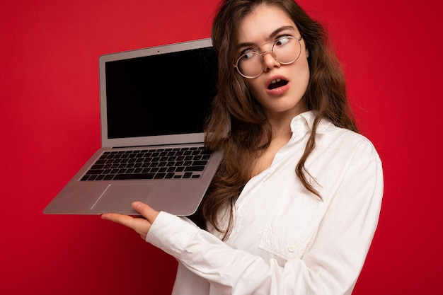 Zdjęcie Dziwne ładnej Brunetki Kręcone Młoda Kobieta Trzyma Komputer Laptop W Okularach Białą Koszulę Patrząc Z Boku Na Wyświetlaczu Na Białym Tle Nad Czerwonym Tle ściany. Skopiuj Przestrzeń, Makiety