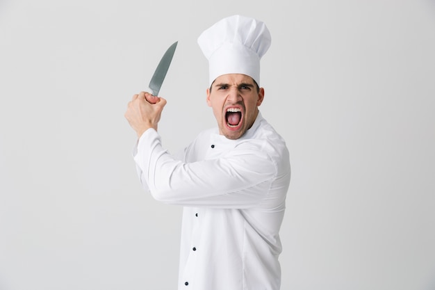 Zdjęcie zdjęcie emocjonalnego agresywnego krzyczącego młodego człowieka szefa kuchni w pomieszczeniu na białym tle na tle białej ściany trzymając nóż.
